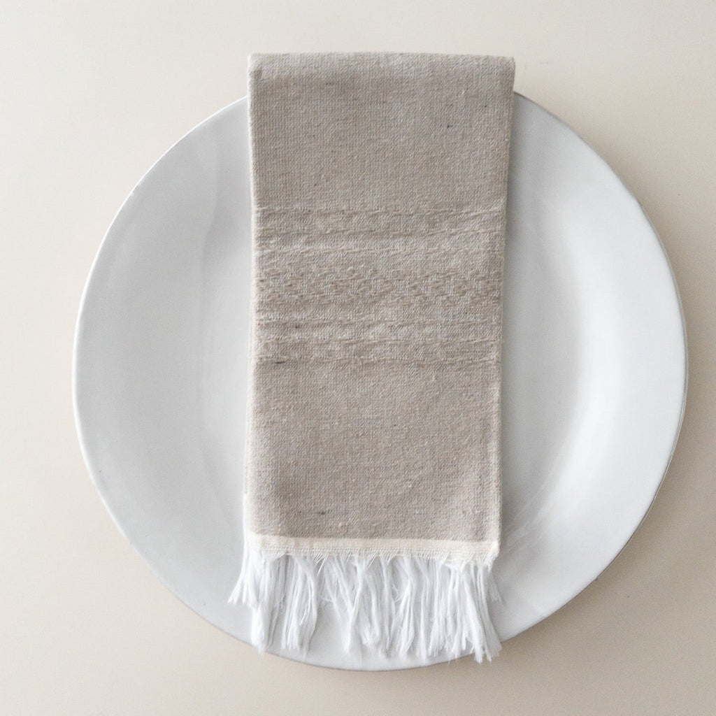 a light gray hand woven napkin with white fringe and a light embroidered pattern on a white glazed ceramic plate