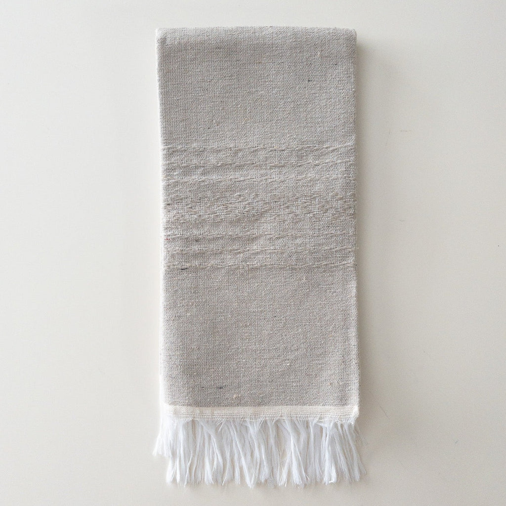 a light gray hand woven napkin with white fringe and a light embroidered pattern