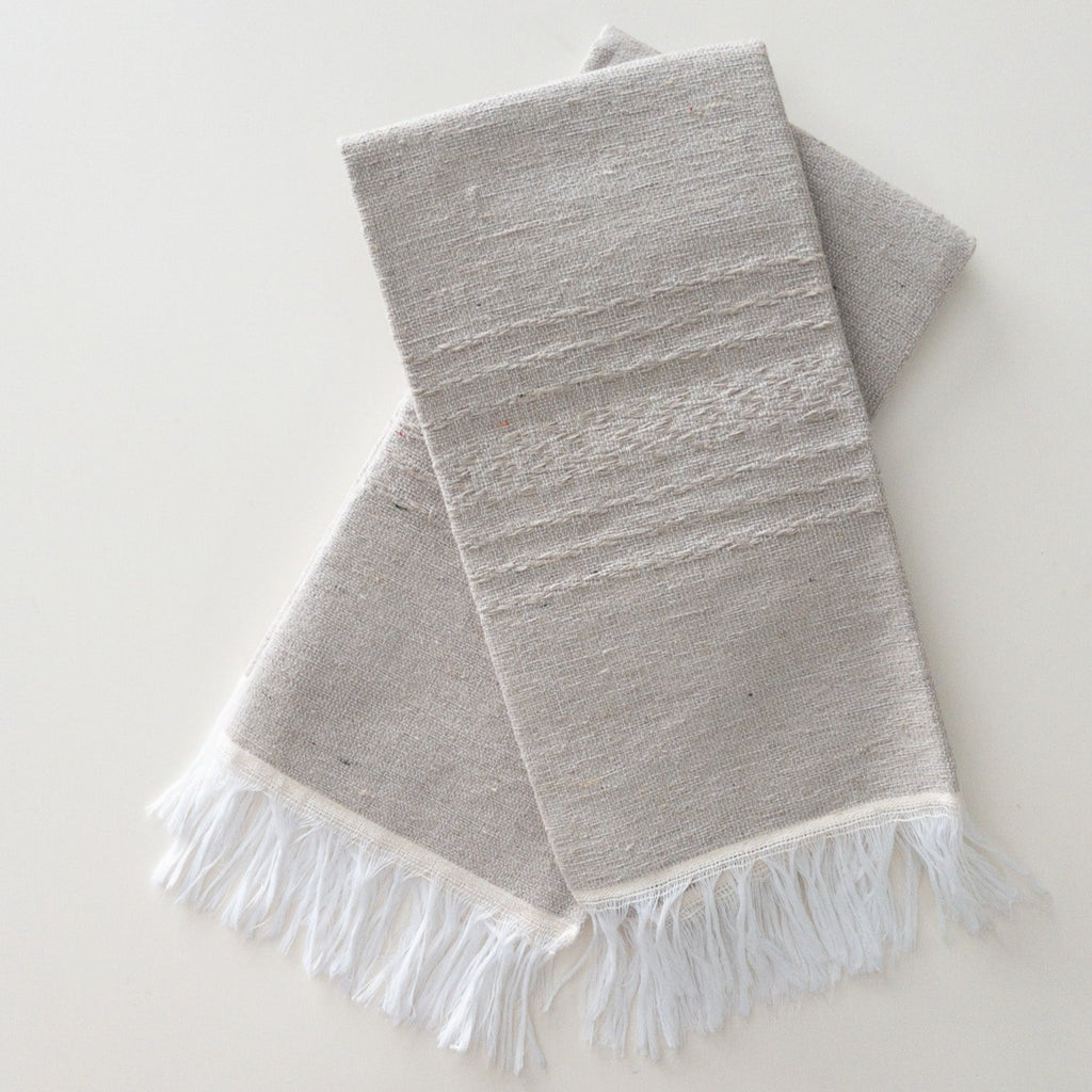 two a light gray hand woven napkins with white fringe and a light embroidered pattern