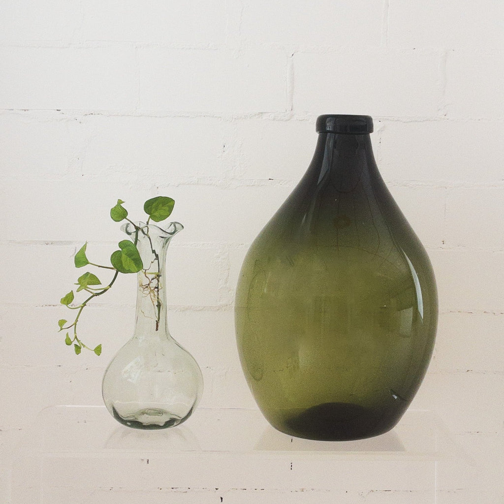 Studio CM | A handblown green vase in a pear shape that is a green with another vase that has a wave top and long neck that is clear holding a small vine