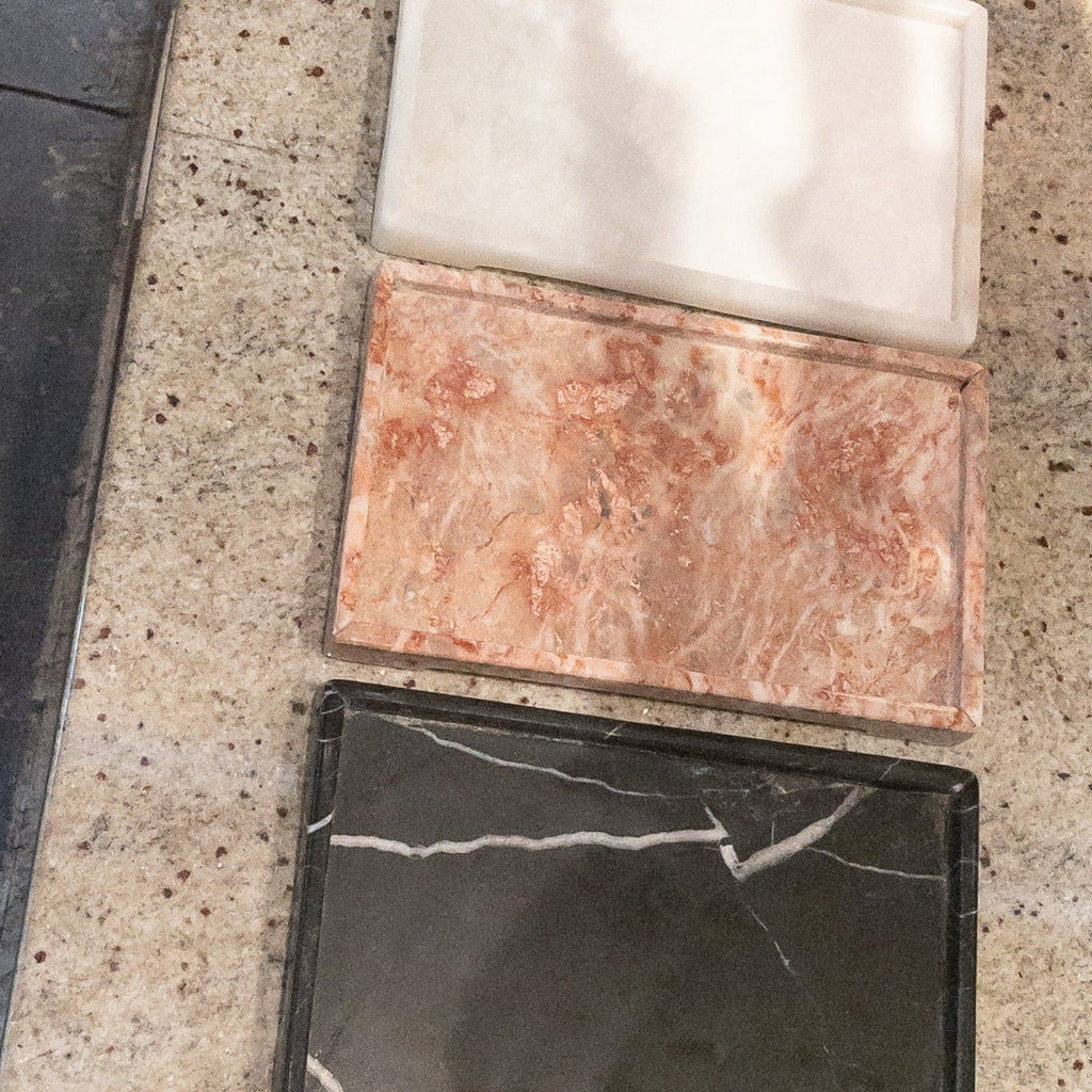 Studio CM | A white onyx tray, pink marble tray and black marble tray sitting side by side on a granite counter top