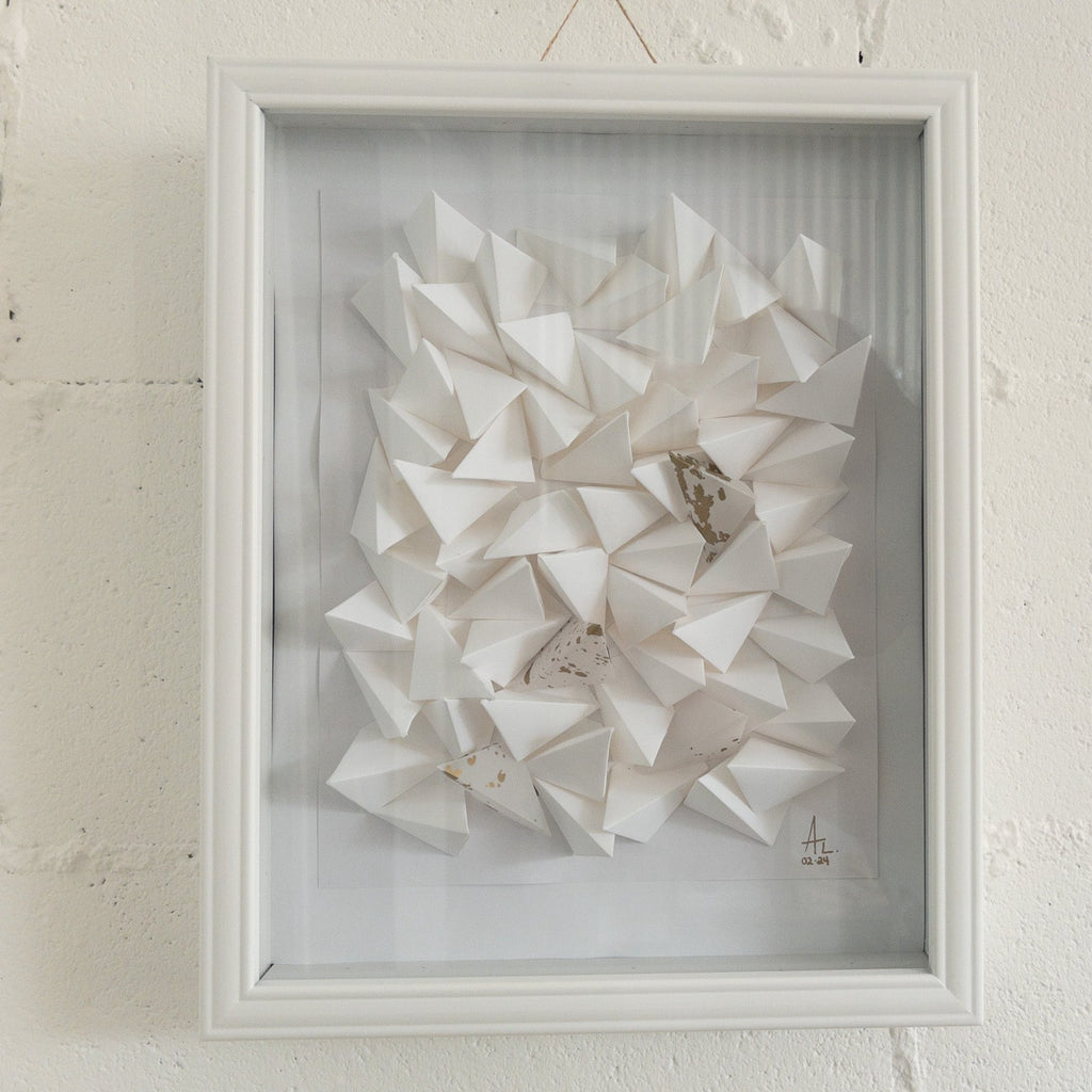 Studio CM | An abstract origami composition framed in a box that has a monotone white color with a touch of gold. Handmade pieces of origami delicately glued together. 