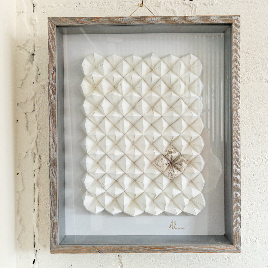 Studio CM | Alicia Lopez art work is hand made origami compositions in a monochrome white with a single corner having a hand painted pattern. Comes framed with glass and light brown wood.