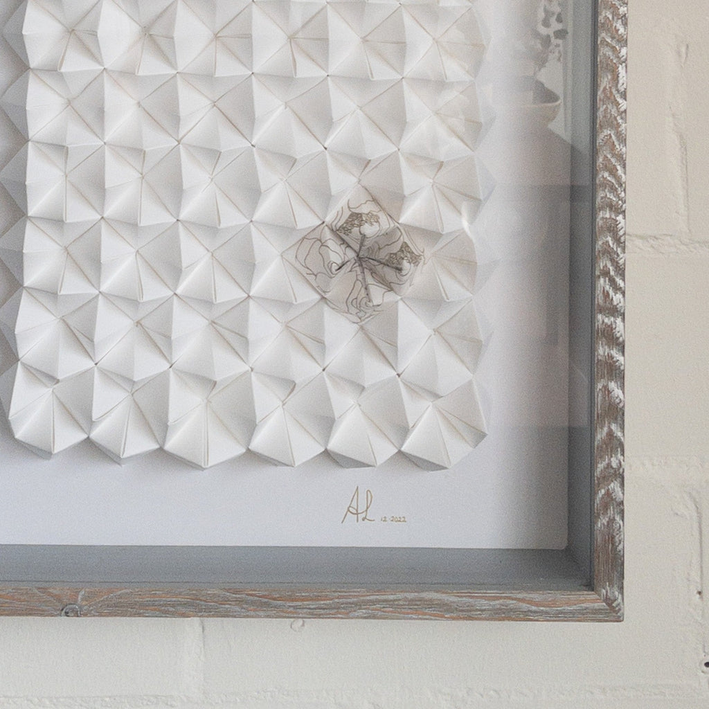 Studio CM | Alicia Lopez art work is hand made origami compositions in a monochrome white with a single corner having a hand painted pattern. Comes framed with glass and light brown wood.