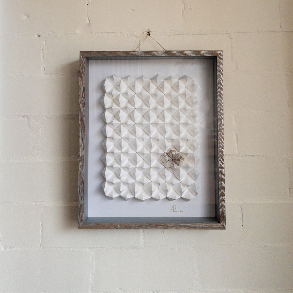 Studio CM | Alicia Lopez art work is hand made origami compositions in a monochrome white with a single corner having a hand painted pattern. Comes framed with glass and light brown wood.