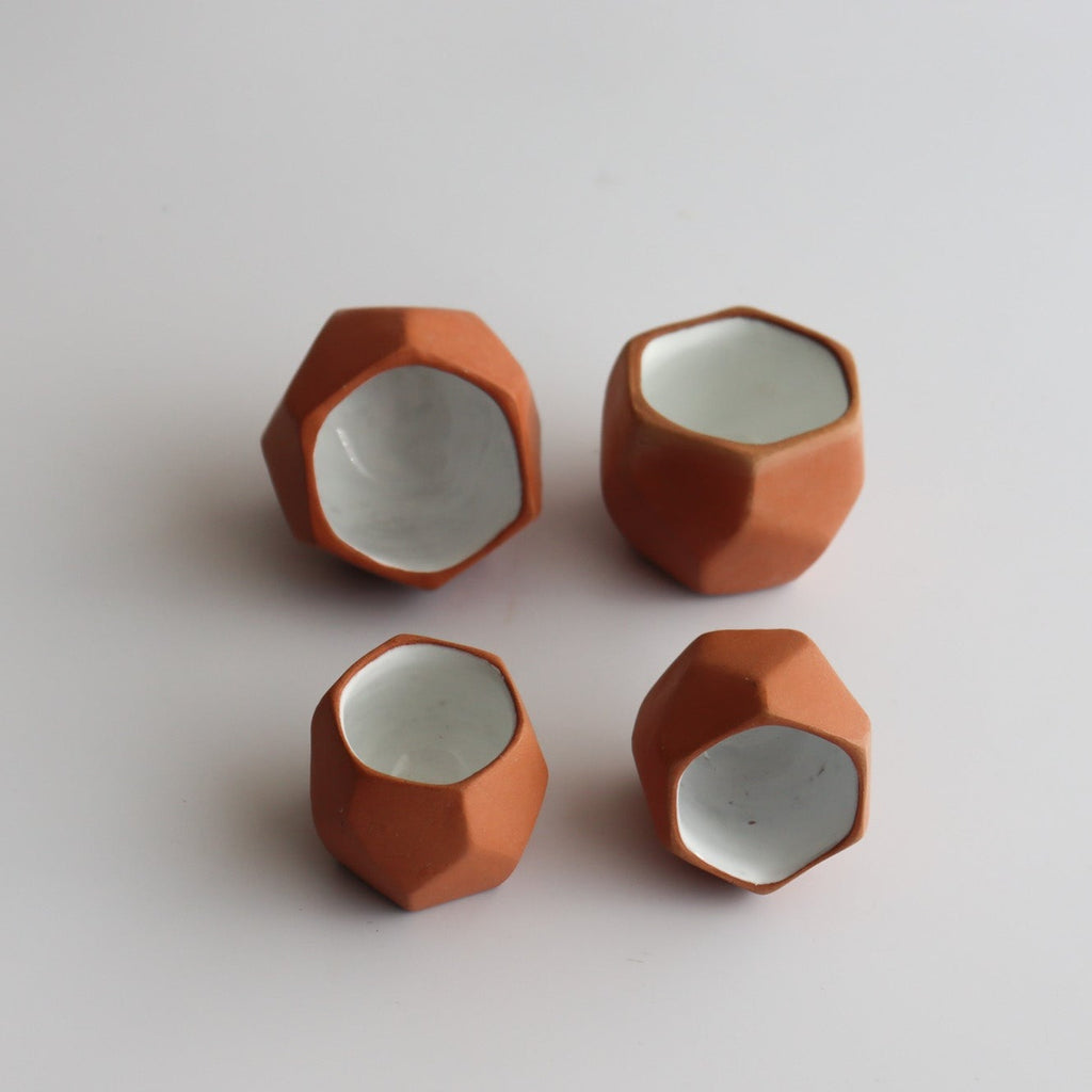 four geometric shaped traditional mexican shot glasses made of clay with a white interior glaze on a white background