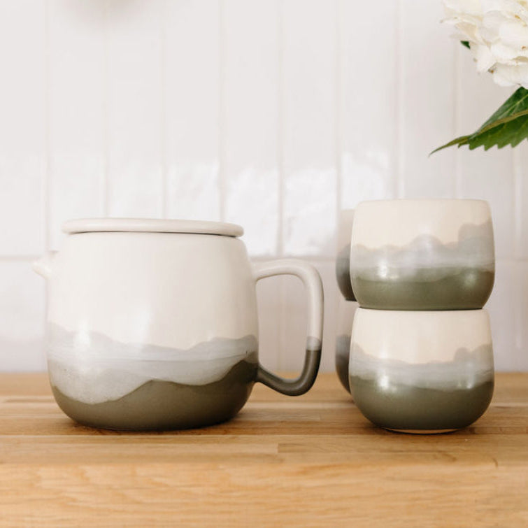 Studio CM | Set of ceramic mugs with gradient design on a wooden surface, next to a vase with flowers.