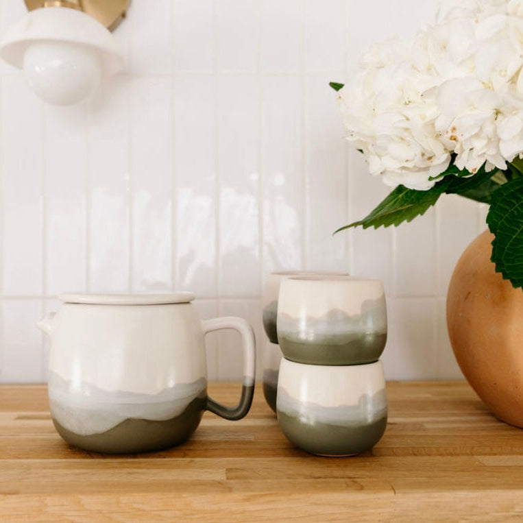 Studio CM | Two ceramic mugs on a wooden surface with a vase of white flowers in the background.