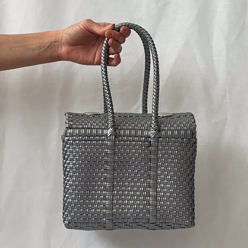 Studio CM | A woven purse made of silver plastic threads in the shape of a box. It has silver metal magnetic buttons to hold it closed and two straps.