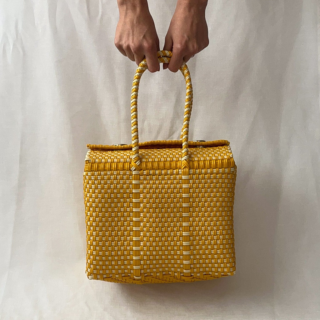 Studio CM | A woven purse made of yellow and white plastic threads in the shape of a box. It has silver metal magnetic buttons to hold it closed and two straps. 