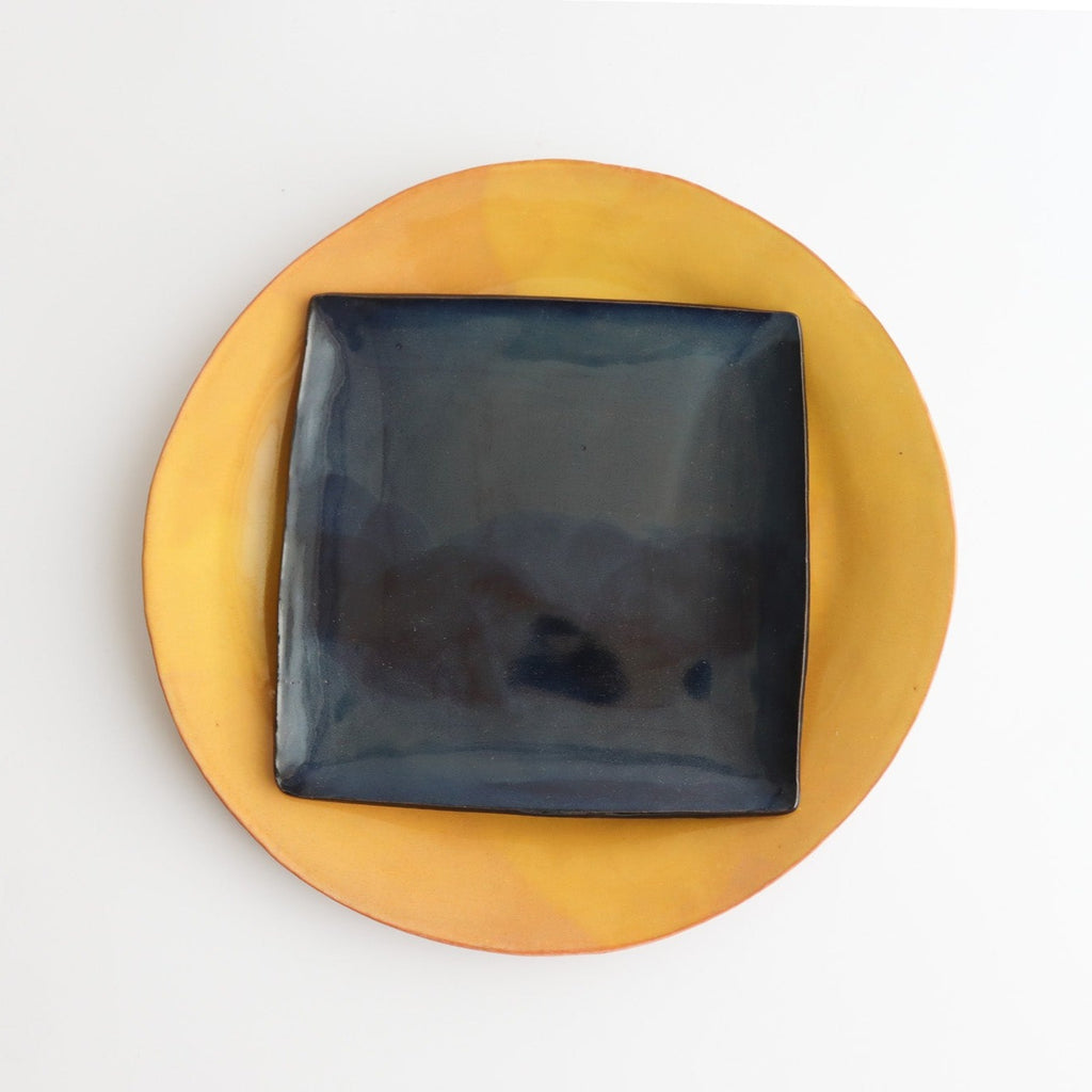 A round yellow ceramic plate with a blue square ceramic plate stacked on top with a white background